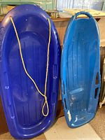 Photo showing two plastic sleds, one dark blue with yellow rope and one light blue with handle.