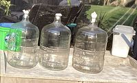 Three large clear glass carboys with stoppers and white plastic container on table in outdoor setting.