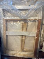 Three wooden canvas frames stacked and wrapped partially in plastic showing stretcher bars and cross braces.