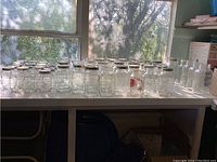 Many clear glass canning jars organized on a white table under a window with natural light. Includes standard round jars and square jars with metal lids, plus smaller bottles without lids.