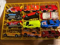 Top-down photo of 11 colorful Majorette die-cast cars arranged in yellow slot holder. Variety of racing and street cars with detailed decals.