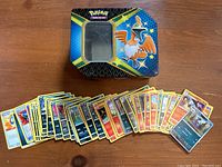 Photo showing Pokémon trading cards spread in a row with collector's tin next to them on a wooden surface.