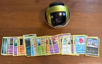 Photo showing a large Ultra Ball collectible container with approximately 50 assorted Pokémon cards laid out in a row beside it on a wooden surface.