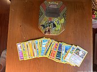 Photo showing the open display of approximately 50 Pokémon cards spread in a fan shape alongside the Pokémon card tin.