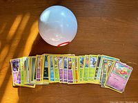 White Premier Ball placed above a spread of approximately 50 Pokémon cards with various types visible, laid out on a wooden surface.