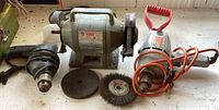 Photo showing a gray King brand bench grinder with two grinding wheels, a corded electric drill with red handle and orange cord, and several grinding and wire brush attachments laid out on floor.
