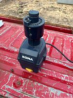 Front view of the black Rona electric drill bit sharpener placed on a red textured surface, showing the adjustable top section with holes for different drill bit sizes and the Rona brand label on the front.