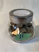 Front view of small glass bottle with metal lid, showing label on lid indicating contents: 60+ pennies, 2 silver dollars, 40+ dimes, 25+ nickels.