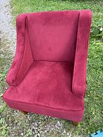 Small red upholstered chair taken outdoors on grass and gravel. Shows the front and side winged backrest with plush upholstery.