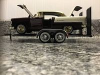 Side view of maroon and white 1955 Chevy Bel Air die-cast model car on black dual-axle trailer showing open hood, door, and trunk.