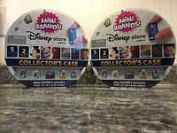 Two sealed 5 Surprise Mini Brands Disney Store Edition Collector's Cases on a countertop with front-facing packaging showing logos, branding, character art, and product details.
