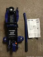 Blue and black 2 ton trolley jack placed on a concrete floor with detached handle and manual next to it.