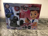 Front view of the boxed children's microscope kit, showing product branding, zoom magnifications (100x, 600x, 1200x), and illustration of a child scientist character.