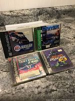 Four PlayStation1 racing game cases shown on granite countertop. Games include NASCAR, Driver, Test Drive Le Mans, and Road Rash 3D. Cases have original cover art and stickers.
