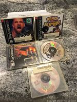 Photo showing 5 PlayStation 1 game discs and cases laid out on a stone surface. Two cases are visible with titles 'Ready 2 Rumble' and 'Brahma Force'. Three discs without cases are also visible.