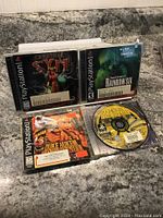 Photo showing three PlayStation 1 game cases and discs: Cardinal Syn, Tom Clancy's Rainbow Six, and Duke Nukem. Cases are open with discs visible.