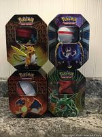 Front view of four closed Pokémon TCG collectible tins featuring Charizard, Raichu, Lunala, and Rayquaza with Poké Ball window on each.