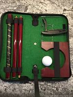 Photo of all set components inside green felt lined black carrying case, displaying disassembled putter, golf ball, and putting cup secured by strap.