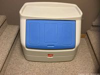 Front view of the Step 2 toy box with blue fold-down lid closed, showing the white plastic body and molded edges, with logo on front.