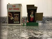 Front view of toy and box on countertop showing outhouse with door open, figure inside, and accessories alongside box with graphics.