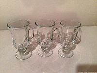 Three clear Irish coffee mugs with pedestal bases and handles, showing their shape and size.