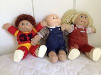 Three original Cabbage Patch dolls sitting on a white surface against a white wall: one in red sportswear with curly brown hair, one bald with overalls, and one with blonde yarn hair in red overalls.