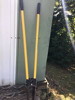 Two manual hole diggers with yellow handles leaning against an outdoor structure, with trees and foliage in the background.