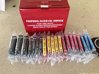 Twenty new Canon ink cartridges arranged side by side, including black (standard and XL), cyan, yellow, and magenta, in sealed plastic sleeves with the original red Canon box behind them.