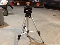 Laser level mounted on aluminum tripod stand with locking leg clips and bubble levels on top, positioned indoors on concrete floor.