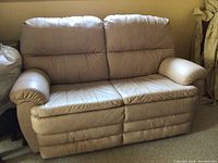Beige leather love seat with two seats, padded arms and cushioned backrest against a wall, shows slight creases from use.
