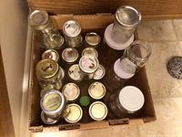 Top view of a cardboard box containing multiple mason jars of varying sizes with metal lids, placed on a tiled floor.