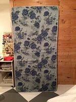 Single bed size foam mattress standing upright against a wooden wall next to a table with sewing materials.