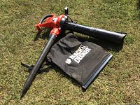 Black and Decker electric leaf blower/mulcher and collection bag laid on grass from side angle, showing handle and tube attachment.
