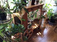 Pine wood tea trolley shown surrounded by plants from two angles. The trolley has a single drawer with brass handles, large spoked wooden wheels on one end, and caster wheels on the other. Top surface shows wear.