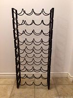 Front view of the wrought iron wine rack showing six shelves with curved bottle holders, black finish, standing against a white wall and floor molding.