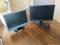 Photo of two computer monitors side by side on a wooden table. Left monitor is a 17-inch Compaq with silver and black casing; right monitor is a 19-inch black LG monitor with power cable coiled on stand.