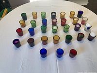 31 votive candle holders of various colors displayed on a white tablecloth. Some holders have candles inside, others are empty.