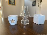 Three candle and tea light holders on a wooden table. One is a white ceramic holder with a star cutout, one is a clear glass hurricane holder, and one is a white square ceramic holder.