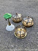 Four ceiling light fixtures including three brass and glass geometric designs and one brass fixture with a green decorative top and frosted white glass shade, displayed on a paved surface.