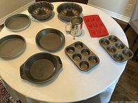 Photo showing 2 bundt cake pans, 2 round cake pans, 2 pie plates, 2 muffin tins, 1 patterned bakeware dish, 1 metal flour sifter, and 1 red silicone baking mold on a white table.