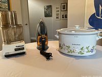 Photo showing three kitchen and household appliances: a Cuisinart food processor, a black and orange steam iron, and a Hamilton Beach crock pot with floral design.