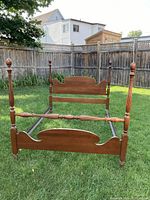 Full view of antique style wooden double bed frame shown outdoors on grass, facing footboard with visible headboard behind, showcasing wood finish and finial posts.