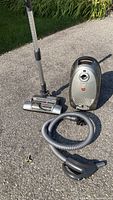Hoover canister vacuum cleaner on asphalt showing hose, telescoping wand, and carpet attachment.