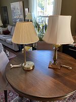 Two table lamps on a wooden table, one with a beige pleated shade and brass base, the other with a cream trapezoidal shade and wooden base with brass.