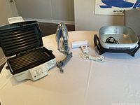 Photo showing electric panini maker, steam iron, travel hair dryer, and electric frying pan with power cord and temperature dial on table.