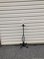 Full view of wrought iron floor candle stick with white taper candle on top, against a beige garage door background.