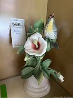 Front view of Boehm porcelain bird statue on white pedestal with large hibiscus flower and green leaves.