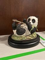 Front left side view of black and white panda figurine lying on back, playing flute, with green scarf and bamboo leaf base on black stand