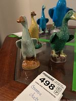 Three ceramic ducks in green, yellow, and light blue on a reflective surface; some show detailed feather texture and bases resembling small patches of terrain