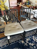 Pair of black bamboo style chairs with beige removable cushions on a blue patterned rug, showing overall wear and stains on cushions.